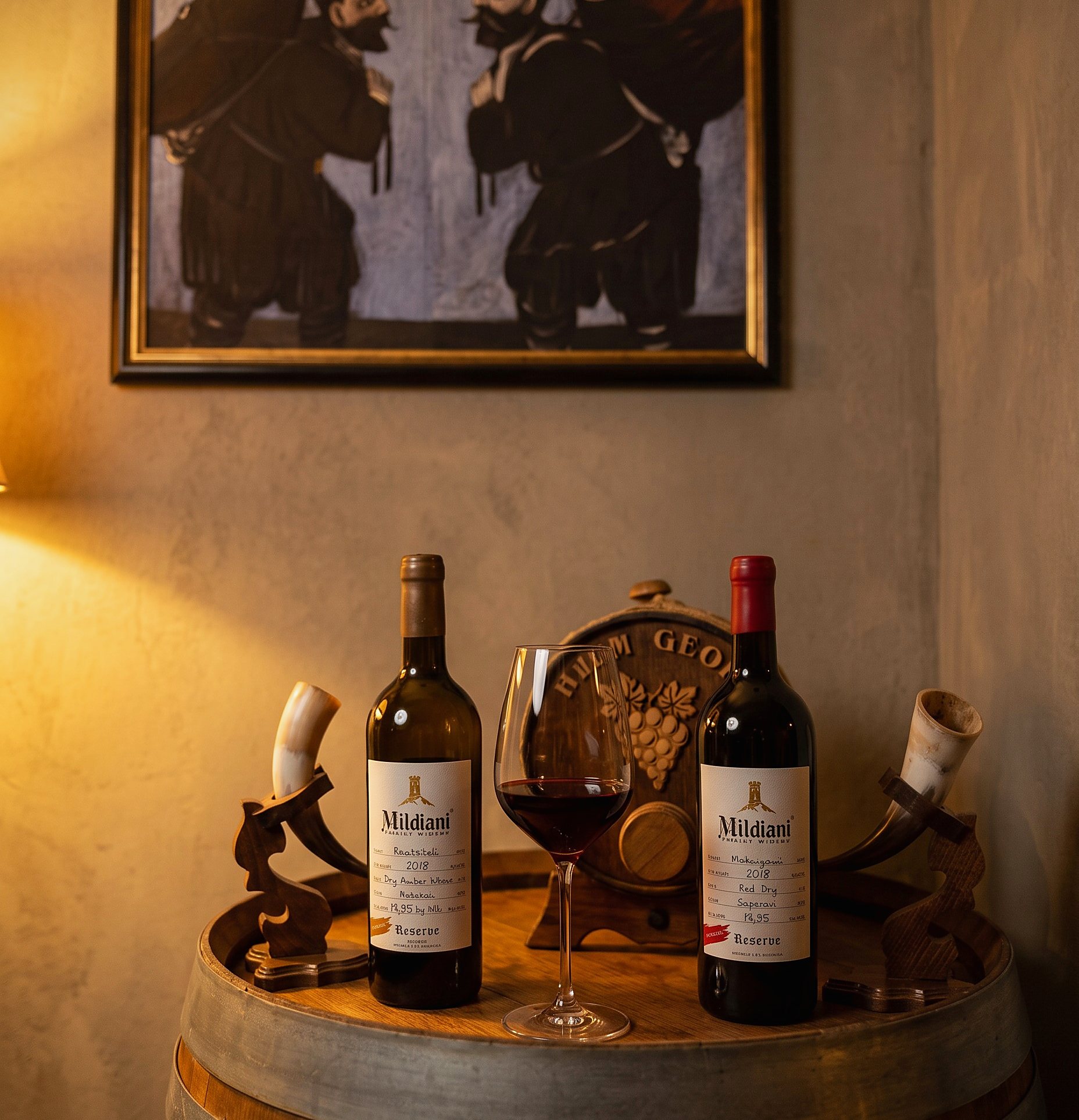 Two bottles of Mildiani Reserve, a poured glass and two drinking horns on the oak barrel, beneath a Georgian painting